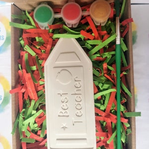 May include: A white ceramic pencil-shaped box with the text "Best Teacher" and a small apple design. The box is in a cardboard box with green and red shredded paper. There are three small paint pots with red, green, and yellow paint and a green paintbrush.