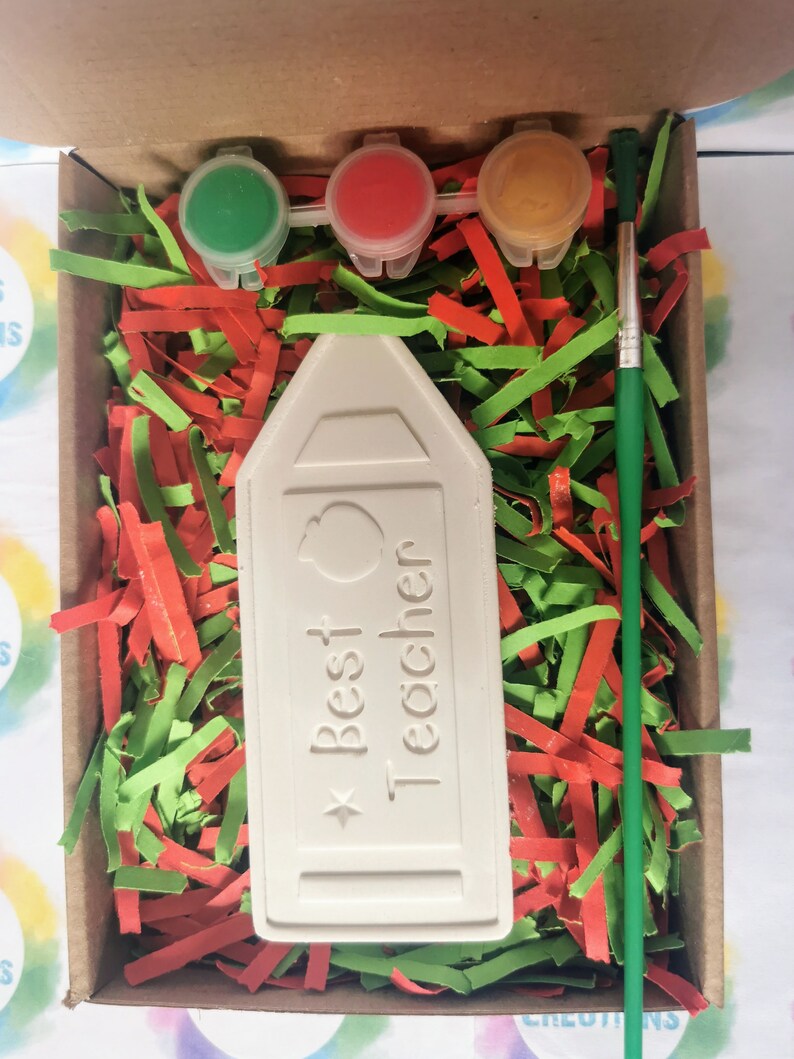 May include: A white ceramic pencil-shaped box with the text "Best Teacher" and a small apple design. The box is in a cardboard box with green and red shredded paper. There are three small paint pots with red, green, and yellow paint and a green paintbrush.