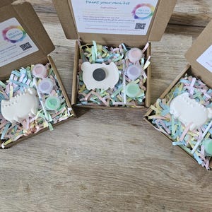 May include: Three open cardboard boxes containing a paint your own kit by Kelly's Creations. Each kit includes a white ceramic cat figure with a magnet on the back, three small paint pots in pink, purple, and green, and a brush, all nestled in pastel paper shreds.