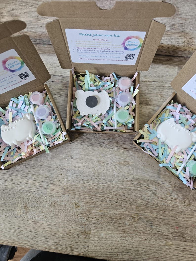May include: Three open cardboard boxes containing a paint your own kit by Kelly's Creations. Each kit includes a white ceramic cat figure with a magnet on the back, three small paint pots in pink, purple, and green, and a brush, all nestled in pastel paper shreds.