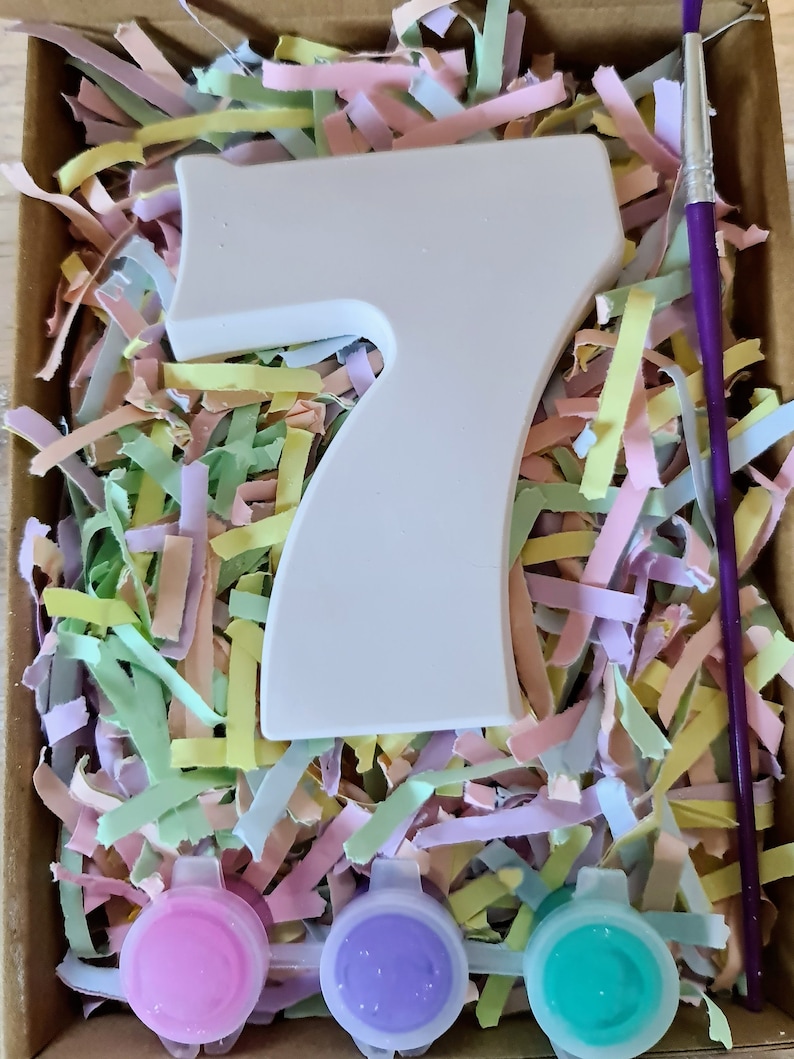 Puede incluir: Un n&uacute;mero siete de cer&aacute;mica blanca, pinceles y tres peque&ntilde;os botes de pintura rosa, morado y turquesa, todo anidado en papel triturado de colores pastel.