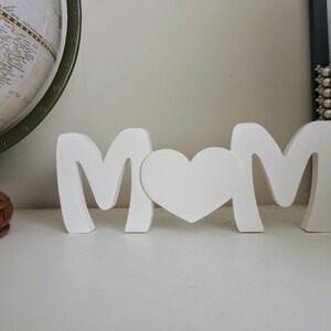 Pu&ograve; includere: Lettere in legno bianco che compongono la parola "MOM" con un cuore bianco tra le lettere. Le lettere sono in piedi su una superficie bianca.