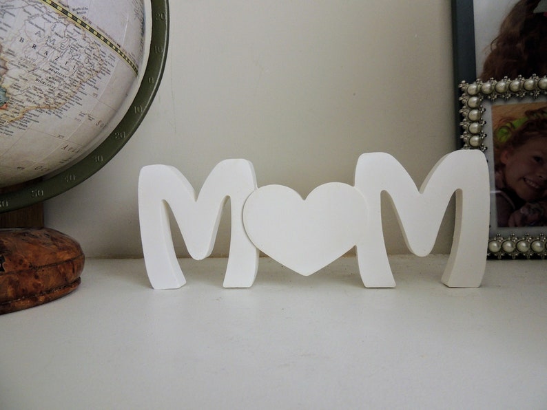 Pu&ograve; includere: Lettere in legno bianco che compongono la parola "MOM" con un cuore bianco tra le lettere. Le lettere sono in piedi su una superficie bianca.