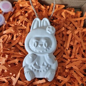 May include: A white ceramic Halloween ornament in the shape of a bunny wearing a costume with a skull and pumpkin design. The ornament is suspended by a twine string and rests on orange paper shreds inside a cardboard box.