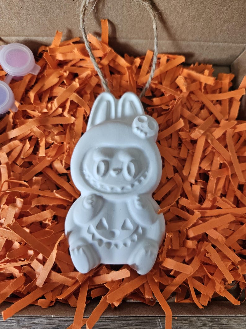 May include: A white ceramic Halloween ornament in the shape of a bunny wearing a costume with a skull and pumpkin design. The ornament is suspended by a twine string and rests on orange paper shreds inside a cardboard box.