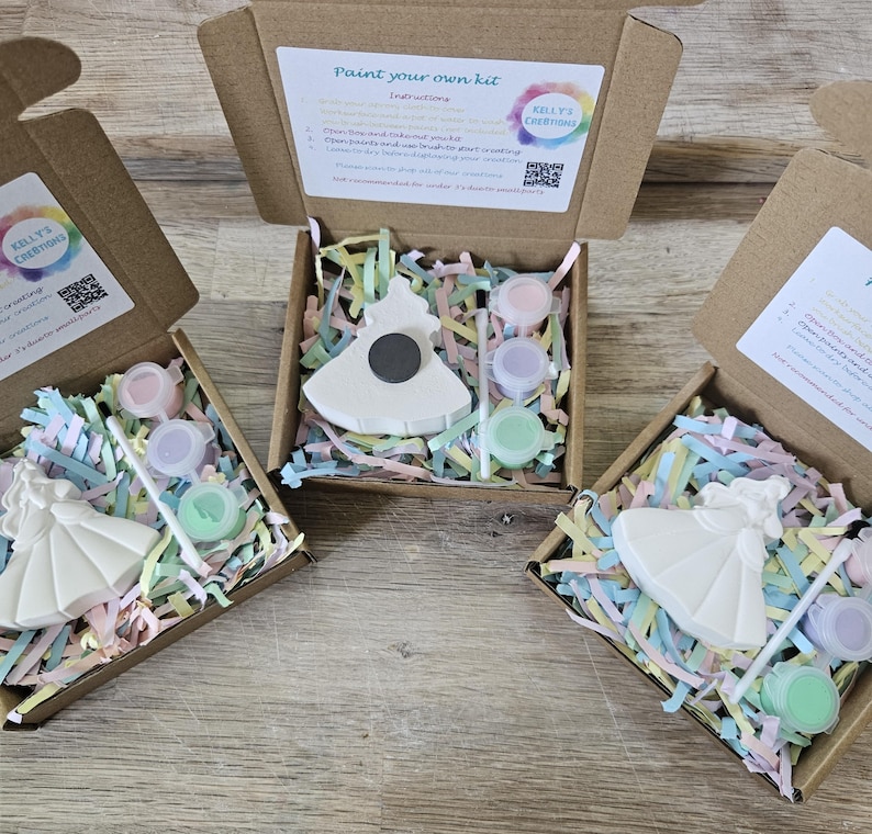 Op de afbeelding: Drie open bruine kartonnen dozen met verfsets. Elke set bevat een wit keramieken beeldje, een klein penseel en drie kleine potjes pastelverf in gekleurde papiersnippers. De deksels van de dozen bevatten instructies.