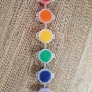 May include: A set of six small, connected paint pots filled with vibrant colors: red, orange, yellow, green, blue, and purple. Each pot has a clear plastic lid and is arranged in a vertical line. The background is a light brown wooden surface.