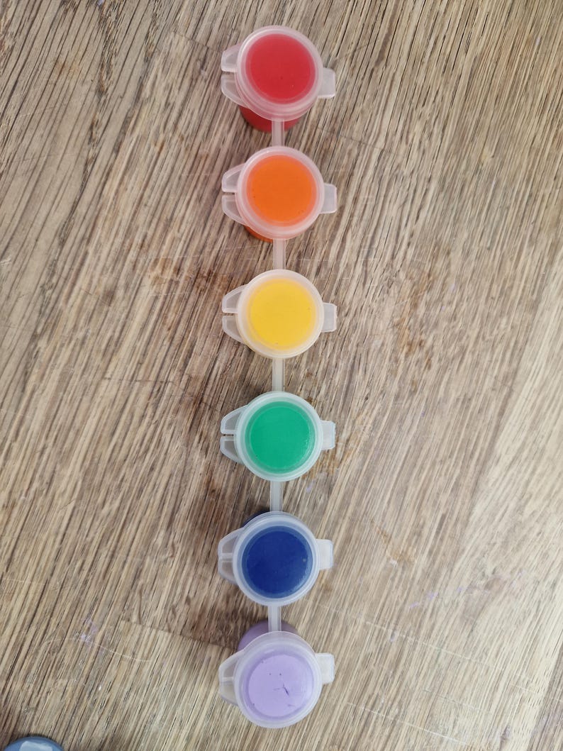 May include: A set of six small, connected paint pots filled with vibrant colors: red, orange, yellow, green, blue, and purple. Each pot has a clear plastic lid and is arranged in a vertical line. The background is a light brown wooden surface.