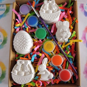May include: A set of six white ceramic Easter-themed figurines, including a bunny, an egg, and a basket, in a box with colorful tissue paper and paint pots.