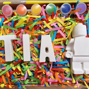 May include: A white ceramic figurine of a person, with the letters "TIA" spelled out in white ceramic letters, surrounded by colorful paper shreds. The figurine and letters are ready to be painted.