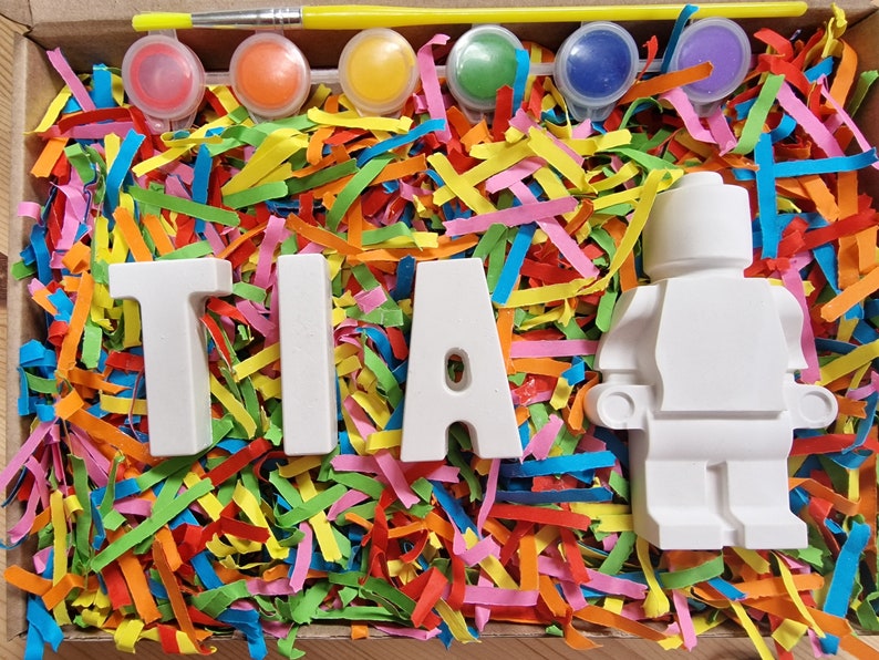 May include: A white ceramic figurine of a person, with the letters "TIA" spelled out in white ceramic letters, surrounded by colorful paper shreds. The figurine and letters are ready to be painted.