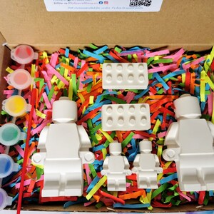 May include: A craft kit with four white unpainted Lego minifigures, two white Lego bricks, and a set of paint pots with brushes. The kit is packaged in a cardboard box with colorful shredded paper.