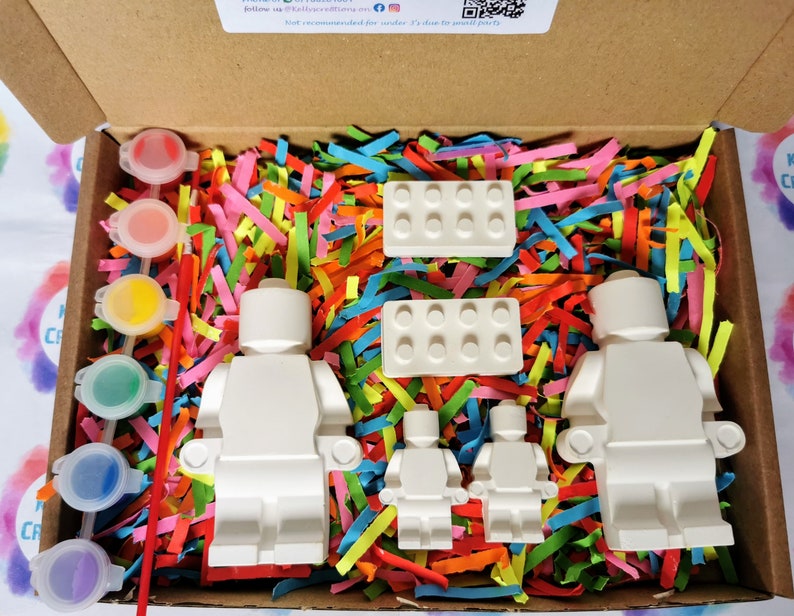 May include: A craft kit with four white unpainted Lego minifigures, two white Lego bricks, and a set of paint pots with brushes. The kit is packaged in a cardboard box with colorful shredded paper.