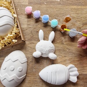 May include: A set of Easter-themed ceramic ornaments, including a bunny, an egg, and a carrot, ready to be painted with a set of colorful paints.