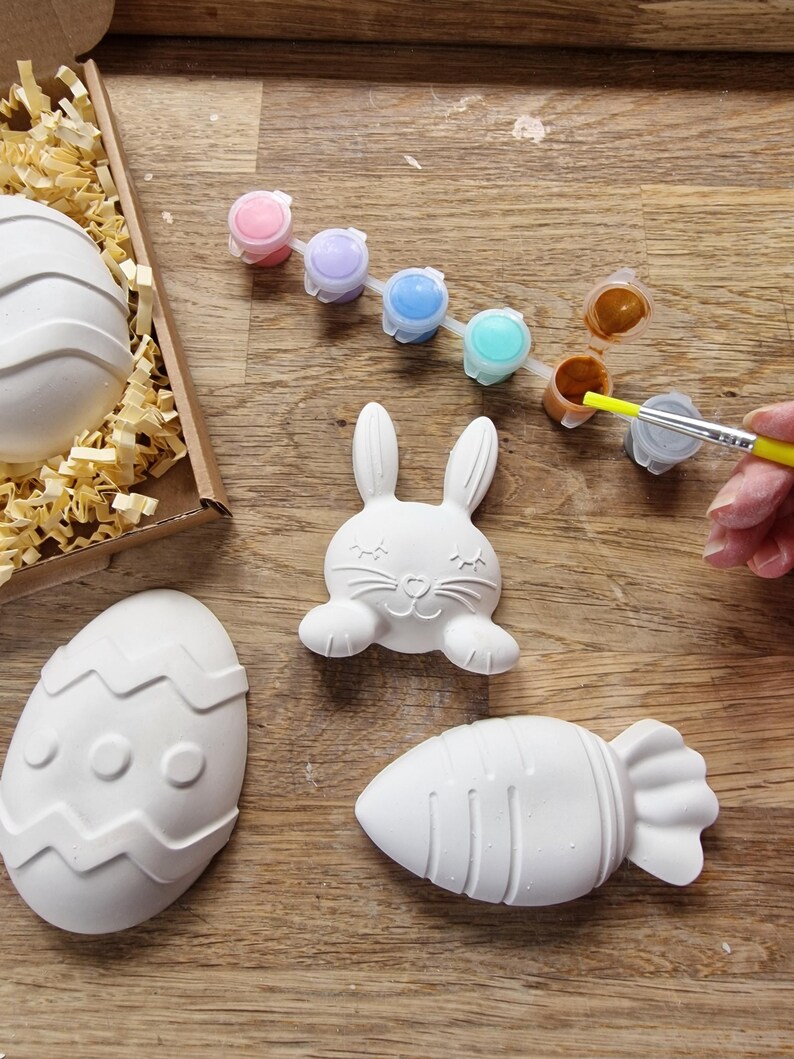 May include: A set of Easter-themed ceramic ornaments, including a bunny, an egg, and a carrot, ready to be painted with a set of colorful paints.