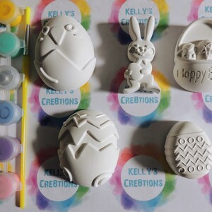 May include: A set of six white ceramic Easter-themed ornaments, including a bunny, a basket, and four eggs, with paint pots and a paintbrush. The ornaments are ready to be painted.