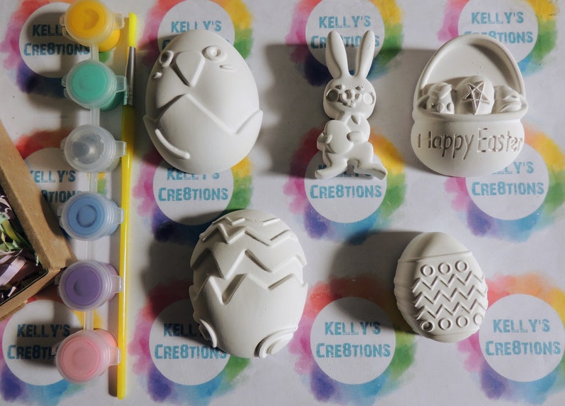 May include: A set of six white ceramic Easter-themed ornaments, including a bunny, a basket, and four eggs, with paint pots and a paintbrush. The ornaments are ready to be painted.