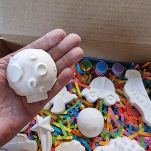 May include: A box filled with white ceramic figurines of various animals, a car, a soccer ball, and a star. The figurines are ready to be painted.
