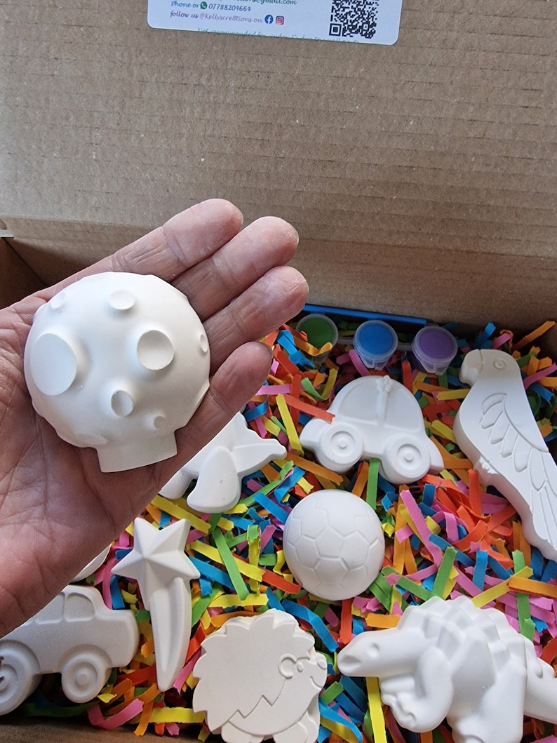May include: A box filled with white ceramic figurines of various animals, a car, a soccer ball, and a star. The figurines are ready to be painted.