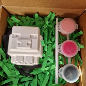May include: A craft kit in a cardboard box, featuring a white block-shaped figure, green paper grass, black paper, three paint pots with pink and gray paint, and a white stirring stick. The text "Not recommended for under 3's due to small parts" is visible.