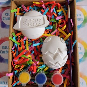 May include: A craft kit for painting ceramic Easter eggs. The kit includes two white ceramic eggs, one with a "Happy Easter" banner, one with a textured design, three paint pots in yellow, blue, and red, and a paintbrush.