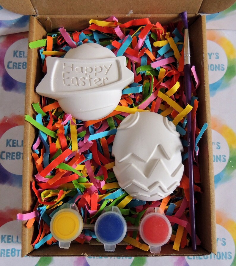 May include: A craft kit for painting ceramic Easter eggs. The kit includes two white ceramic eggs, one with a "Happy Easter" banner, one with a textured design, three paint pots in yellow, blue, and red, and a paintbrush.