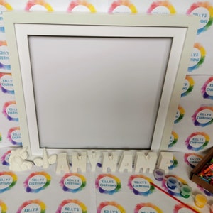 May include: A white wooden frame with a blank canvas inside.  The frame is surrounded by colorful rainbow circles with the text "Kelly's Creations" on each circle.  Below the frame are white ceramic letters spelling out "ANYNAME" and small paint pots with paintbrushes.