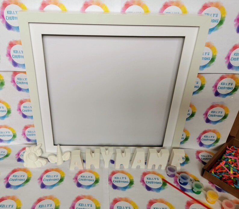 May include: A white wooden frame with a blank canvas inside.  The frame is surrounded by colorful rainbow circles with the text "Kelly's Creations" on each circle.  Below the frame are white ceramic letters spelling out "ANYNAME" and small paint pots with paintbrushes.