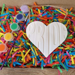 Puede incluir: Un kit de manualidades en una caja de cart&oacute;n llena de papel triturado de colores. El kit incluye un objeto blanco en forma de coraz&oacute;n con la palabra "PRIDE" y peque&ntilde;os botes de pintura con un pincel. Los colores son morado, azul, verde, amarillo, naranja y rojo.