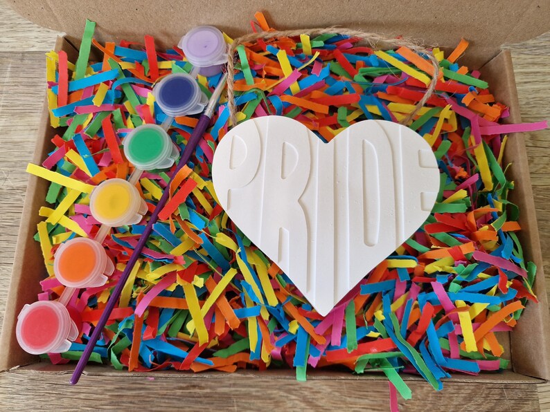 Puede incluir: Un kit de manualidades en una caja de cart&oacute;n llena de papel triturado de colores. El kit incluye un objeto blanco en forma de coraz&oacute;n con la palabra "PRIDE" y peque&ntilde;os botes de pintura con un pincel. Los colores son morado, azul, verde, amarillo, naranja y rojo.