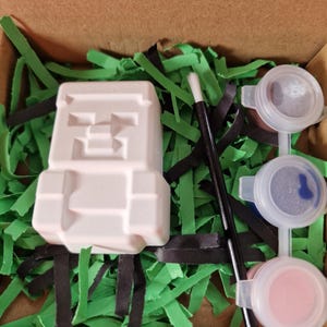 May include: A white, Minecraft-themed plaster figure, paint pots with pink and blue paint, and a paintbrush are arranged in a cardboard box with green and black paper grass. The figure has a pixelated design.