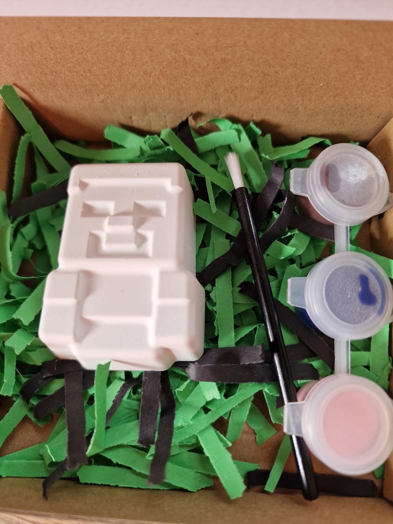 May include: A white, Minecraft-themed plaster figure, paint pots with pink and blue paint, and a paintbrush are arranged in a cardboard box with green and black paper grass. The figure has a pixelated design.