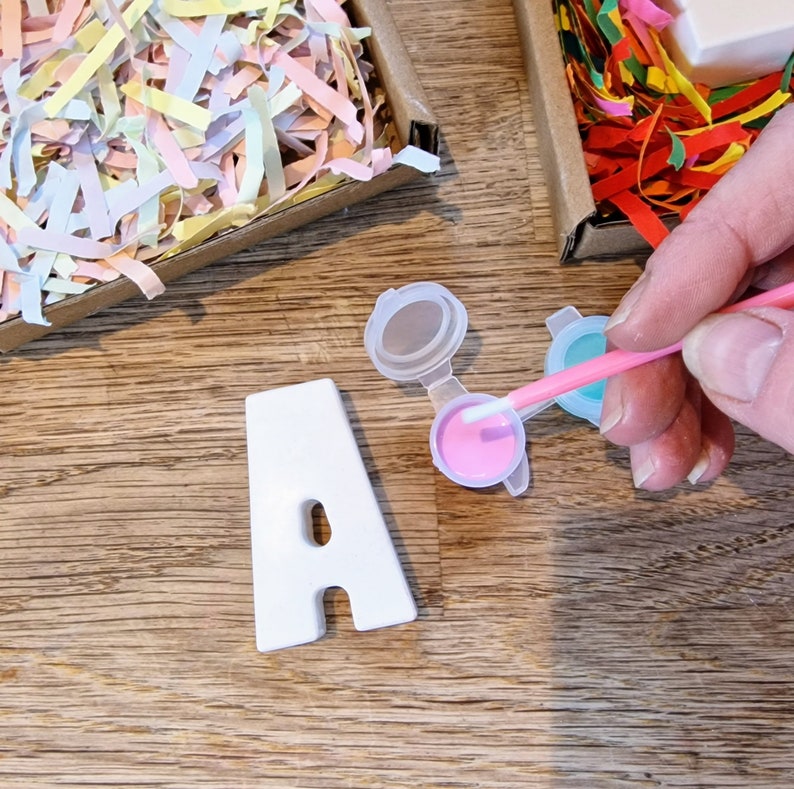 May include: A white ceramic letter A with a hole in the middle, being painted pink with a small paintbrush. The letter is on a wooden surface with two small containers of paint, one pink and one blue.