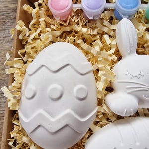 May include: A craft kit with white ceramic Easter egg, bunny, and carrot shapes, ready to be painted. The kit includes four small bottles of paint in pink, purple, blue, and green.