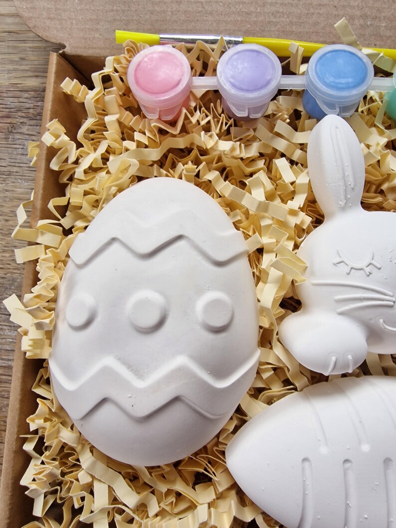 May include: A craft kit with white ceramic Easter egg, bunny, and carrot shapes, ready to be painted. The kit includes four small bottles of paint in pink, purple, blue, and green.
