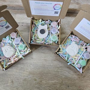 Puede incluir: Tres cajas de cart&oacute;n marr&oacute;n abiertas que contienen kits de pintura. Cada kit incluye una figura de yeso blanco con forma de gato, un pincel peque&ntilde;o y tres recipientes circulares con pintura pastel sobre papel triturado de colores.