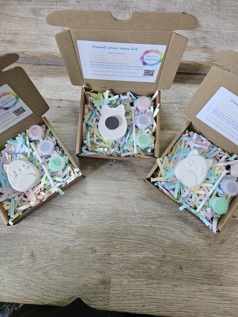 Puede incluir: Tres cajas de cart&oacute;n marr&oacute;n abiertas que contienen kits de pintura. Cada kit incluye una figura de yeso blanco con forma de gato, un pincel peque&ntilde;o y tres recipientes circulares con pintura pastel sobre papel triturado de colores.
