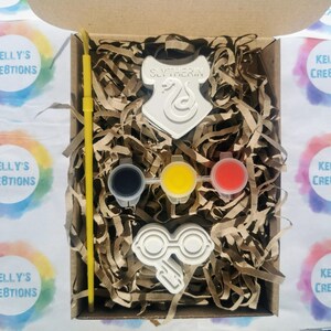 May include: A craft kit in a cardboard box, featuring a white Slytherin crest, glasses, and three paint pots with black, yellow, and red paint. A yellow paintbrush and shredded paper fill the box. The box is labeled "Kelly's Creations".