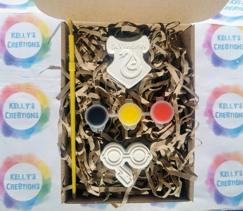 May include: A craft kit in a cardboard box, featuring a white Slytherin crest, glasses, and three paint pots with black, yellow, and red paint. A yellow paintbrush and shredded paper fill the box. The box is labeled "Kelly's Creations".