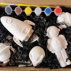 May include: A craft kit containing five white ceramic figurines: a rocket, an astronaut, an alien, and two planets. The kit includes six small pots of paint in various colours.