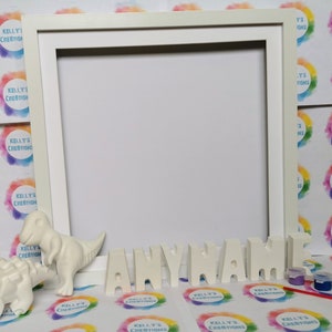 May include: A white picture frame with a blank space, alongside ceramic dinosaur figures and the word "ANYNAME" in individual letters. Small paint pots and a paintbrush are included, indicating a DIY craft kit.