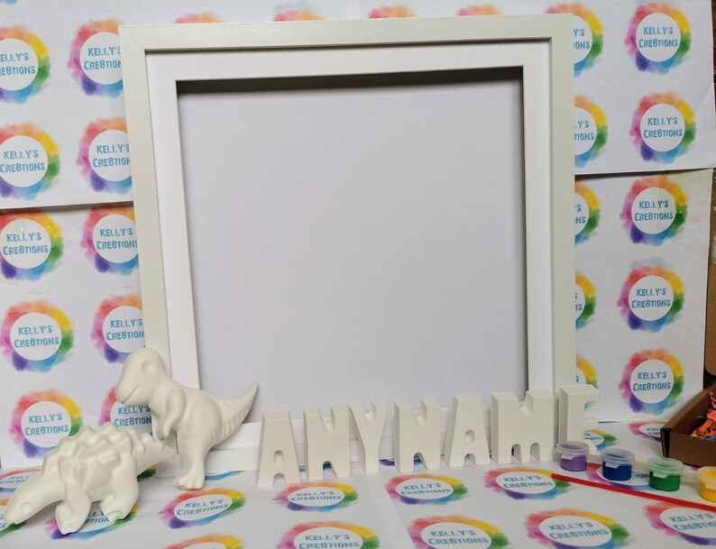 May include: A white picture frame with a blank space, alongside ceramic dinosaur figures and the word "ANYNAME" in individual letters. Small paint pots and a paintbrush are included, indicating a DIY craft kit.