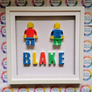 May include: A framed piece of art with two Lego-style figures in red and blue, and the name "BLAKE" in colourful letters. The artwork is set against a white background within a white frame. The background includes the text "KELLY'S CREATIONS".