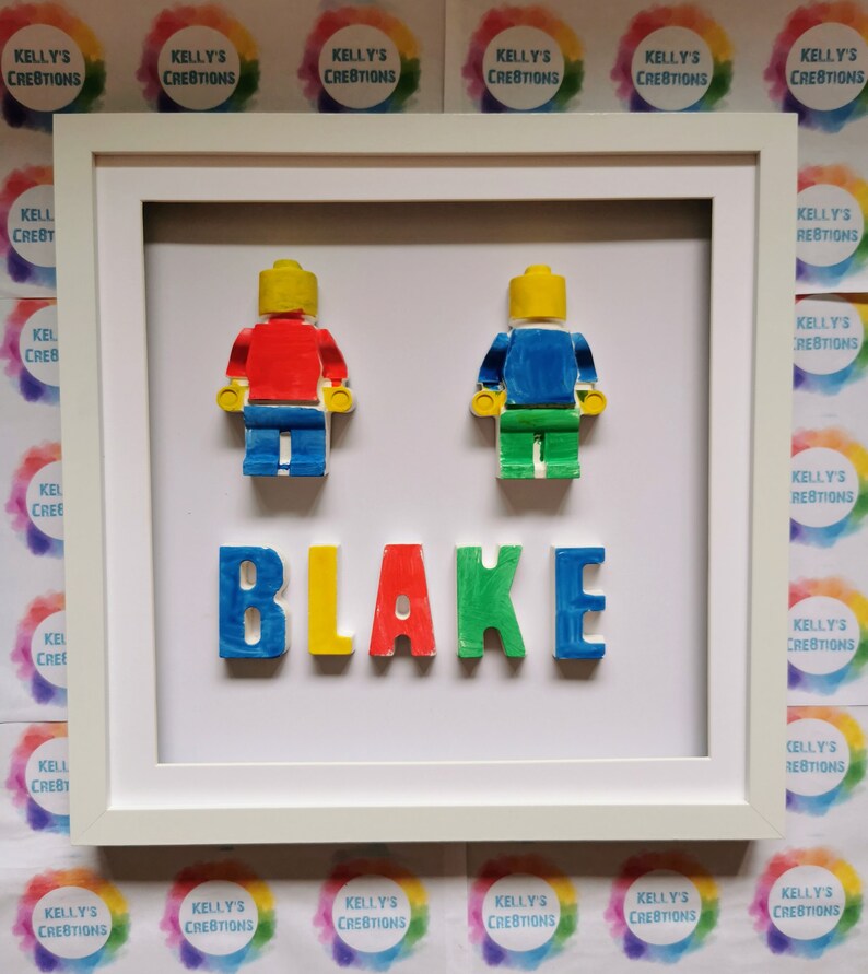 May include: A framed piece of art with two Lego-style figures in red and blue, and the name "BLAKE" in colourful letters. The artwork is set against a white background within a white frame. The background includes the text "KELLY'S CREATIONS".