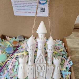 May include: A white ceramic castle ornament suspended by twine, part of a paint-your-own kit. The kit includes paint pots and instructions. The castle has multiple turrets and a detailed facade, resting on colorful shredded paper inside a cardboard box.