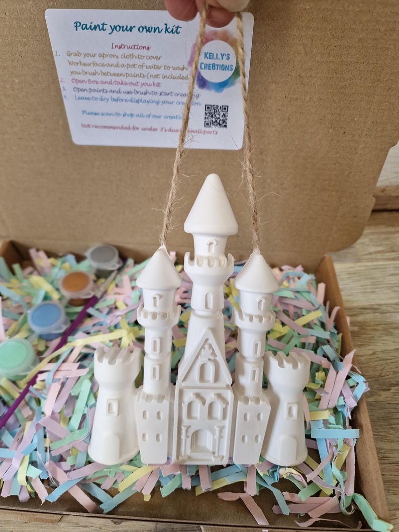 May include: A white ceramic castle ornament suspended by twine, part of a paint-your-own kit. The kit includes paint pots and instructions. The castle has multiple turrets and a detailed facade, resting on colorful shredded paper inside a cardboard box.
