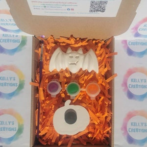 May include: A Halloween-themed craft kit in a cardboard box. The kit includes a white bat and pumpkin, three paint pots, and a paintbrush. The box is filled with orange paper shreds. The kit is from Kelly's Creations.
