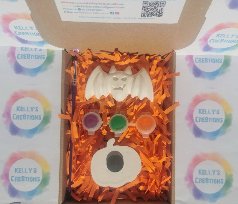 May include: A Halloween-themed craft kit in a cardboard box. The kit includes a white bat and pumpkin, three paint pots, and a paintbrush. The box is filled with orange paper shreds. The kit is from Kelly's Creations.
