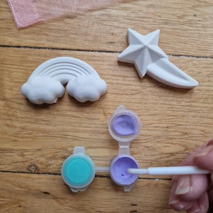 Puede incluir: Un kit de manualidades con tres piezas de cer&aacute;mica blanca: un arco&iacute;ris, una estrella y una estrella fugaz. El kit incluye dos peque&ntilde;os botes de pintura, uno con pintura morada y otro con pintura turquesa. Se muestra un pincel sumergido en la pintura morada.