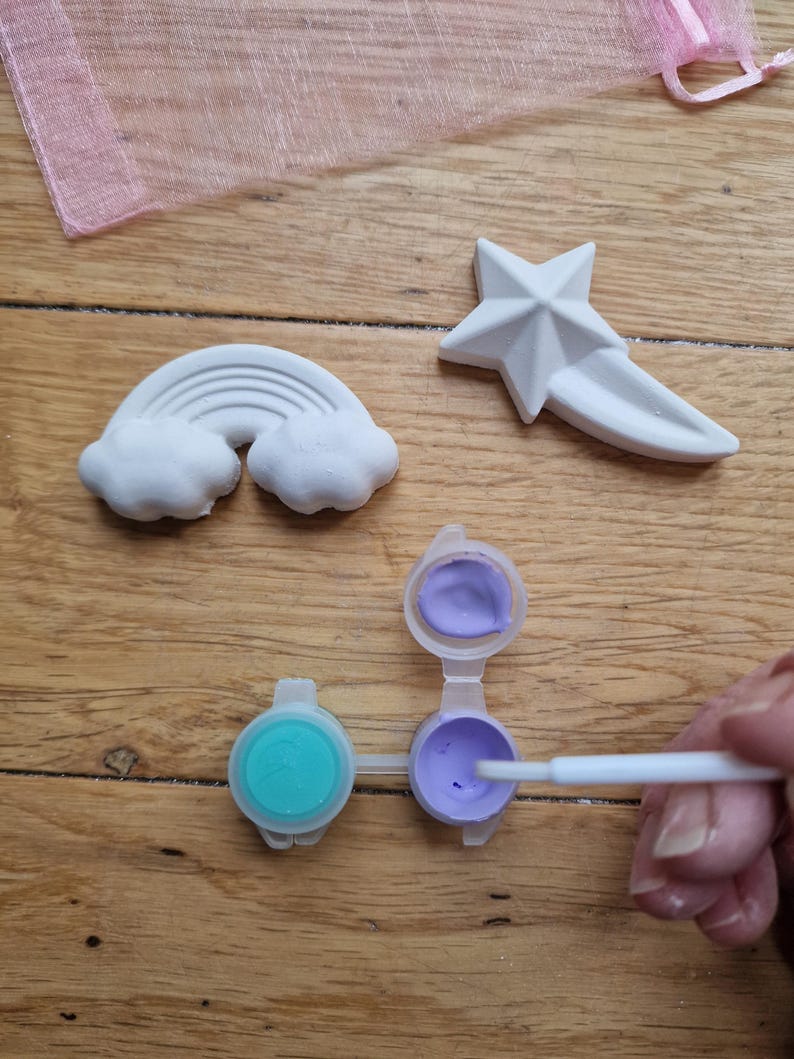 Puede incluir: Un kit de manualidades con tres piezas de cer&aacute;mica blanca: un arco&iacute;ris, una estrella y una estrella fugaz. El kit incluye dos peque&ntilde;os botes de pintura, uno con pintura morada y otro con pintura turquesa. Se muestra un pincel sumergido en la pintura morada.